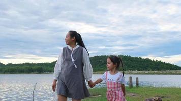 la madre felice cammina mano nella mano di una bambina sorridente che si gode la natura insieme in riva al lago. famiglia felice, madre e figlia che giocano sentendosi felici nel parco. video