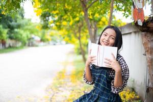 niña sentada en un banco leyendo un libro bajo un hermoso árbol. foto