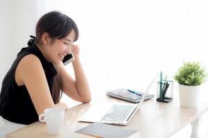 hermosa joven mujer asiática independiente sonriendo trabajando en una computadora portátil y hablando de un teléfono inteligente móvil en la oficina de escritorio con una chica profesional que usa el concepto de cuaderno, negocios y estilo de vida. foto