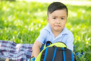 niño asiático joven que sonríe y que abre la mochila en el césped, expresión linda del niño de asia divertido y bolso de escuela abierto feliz, concepto de la educación. foto