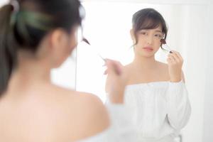 Beauty portrait young asian woman smiling with face looking mirror applying makeup with brush cheek in the bedroom, beautiful girl holding blusher, skin care and cosmetic table fashion concept. photo