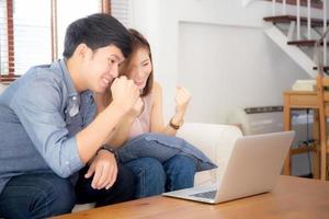 Beautiful portrait asian young couple working laptop with smile and happy sitting on couch at living room, man and woman on sofa using notebook computer with excited, business and success concept. photo