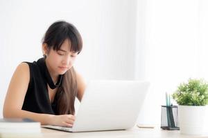 Young freelance asian woman smiling working and typing on laptop computer at desk office with professional, girl using notebook checking email or social network, business and lifestyle concept. photo