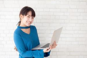 Beautiful portrait asian young woman smile using laptop standing at workplace on cement concrete background, girl happy with computer internet online, lifestyle and freelance business concept. photo