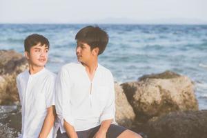 Homosexual portrait young asian couple sitting hug together on rock or stone in the beach in summer, asia gay going tourism for leisure and relax with happy in vacation at sea, LGBT legal concept. photo