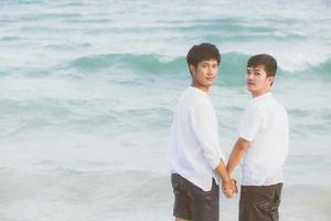 Back view homosexual asian couple standing together on beach in summer, asia gay holding hands going sea for leisure and relax with romantic and happy in vacation at sea, LGBT with legal concept. photo