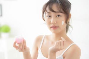 beautiful young asian woman happy applying cream or lotion with moisturizer to skin face, beauty asia girl applying skincare touch facial with cosmetic makeup, healthy and wellness concept. photo