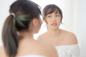 Beauty portrait young asian woman smiling look at mirror of checking skin care caucasian with wellness in the bedroom, beautiful girl happy touching face in reflection for health, lifestyle concept. photo