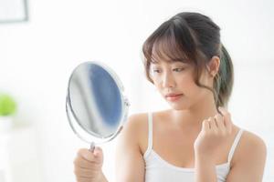 Beautiful young asian woman looking the mirror with acne problem at bedroom, trouble of beauty on face, zit treatment, asia girl is pimple having worry and displeased, skincare and healthy concept. photo