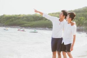 Homosexual portrait young asian couple standing pointing something together on beach in summer, asia gay going sea for leisure with romantic and happy in vacation at sea, LGBT with legal concept. photo