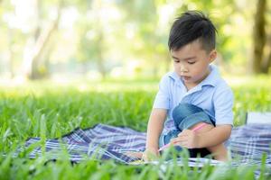 Young little asian boy reading and writing book in the park, asia kid homework and study in the summer, child relax with drawing on notebook in vacation, education and development concept. photo
