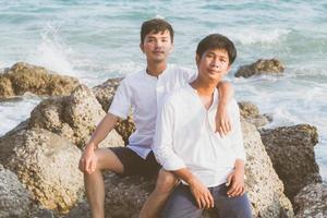 retrato homosexual joven pareja asiática sentada abrazada en roca o piedra en la playa en verano, turismo gay de asia para el ocio y relajarse con felicidad en vacaciones en el mar, concepto legal lgbt. foto