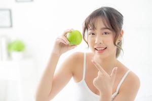 Beautiful portrait young asian woman holding and pointing green apple fruit in the bedroom at home, lifestyle of nutrition girl healthy and care weight loss, health and wellness concept. photo