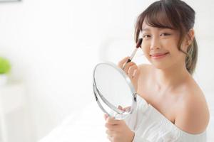 Beauty portrait young asian woman smile with face looking mirror applying makeup with brush cheek in the bedroom, beautiful of girl holding blusher, skin care and cosmetic concept. photo