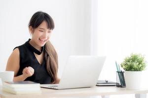 hermoso retrato mujer joven asiática sonríe trabajando en una computadora portátil en línea con feliz satisfecha sentada en el escritorio de la oficina, chica usando un cuaderno con el concepto emocionado y alegre, de negocios y de éxito. foto