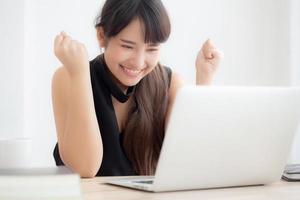 Beautiful portrait asian young woman smile working online laptop computer with happy satisfied sitting on desk office, girl using notebook with excited and glad, business and success concept. photo