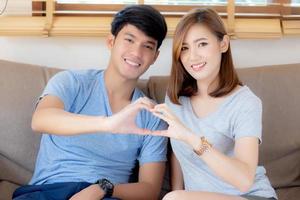 Beautiful happy young couple sitting on sofa fun making gesture heart shape with hand indoor together, man and woman relation feeling love with symbol and sign, lover and romantic concept. photo