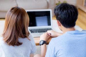 Back view of young asian couple working laptop with blank screen display, family planning and searching content together, man and woman looking computer, business and communication concept. photo