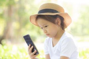 hermoso niño asiático sonriendo usando un teléfono móvil inteligente en el jardín, el niño tiene pasión por jugar juegos digitales en un teléfono inteligente en la red de Internet en el parque con un concepto feliz, de estilo de vida y tecnológico. foto