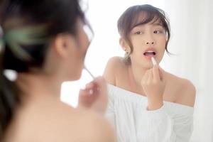 hermoso retrato joven mujer asiática mirando espejo aplicando lápiz labial de maquillaje en la habitación, labios de belleza maquillaje de niña de asia y moda cosmética en la boca en casa, estilo de vida y concepto de atención médica. foto