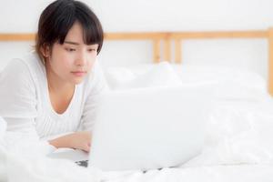Beautiful portrait asian young woman smile lying on bed using laptop computer with happy at bedroom for leisure and relax, freelance with girl working notebook, communication concept. photo
