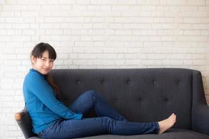 hermoso retrato joven mujer asiática sentada y sonriendo feliz y mirando la cámara en el sofá con informal en la sala de estar, chica alegre y relajada en el sofá en casa, concepto de estilo de vida. foto