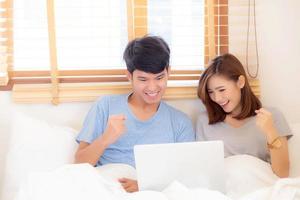 Beautiful portrait young asian couple working laptop with smile and happy sitting in bedroom, man and woman using notebook computer with excited and satisfied, business and success concept. photo