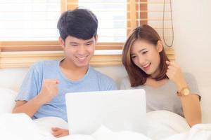 Beautiful portrait young asian couple working laptop with smile and happy sitting in bedroom, man and woman using notebook computer with excited and satisfied, business and success concept. photo