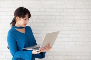 Beautiful portrait asian young woman smile using laptop standing at workplace on cement concrete background, girl happy with computer internet online, lifestyle and freelance business concept. photo