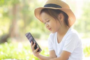 hermoso niño asiático sonriendo usando un teléfono móvil inteligente en el jardín, el niño tiene pasión por jugar juegos digitales en un teléfono inteligente en la red de Internet en el parque con un concepto feliz, de estilo de vida y tecnológico. foto