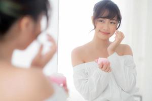 Beautiful portrait young asian woman applying cream moisturizer or lotion skin care cosmetic on face looking mirror, girl with treatment facial, health and wellness concept. photo