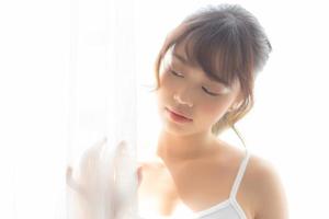 retrato de una hermosa joven asiática parada en la ventana y sonriendo mientras se despierta con el amanecer en la mañana, una chica feliz con frescura y alegre, estilo de vida y concepto de relajación. foto