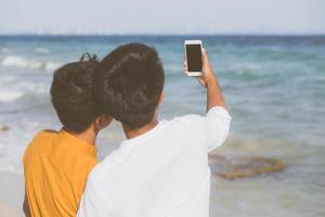 Back view gay portrait young couple smiling taking a selfie photo together with smart mobile phone at beach, LGBT homosexual lover in the vacation at sea, two man going to travel, holiday concept.