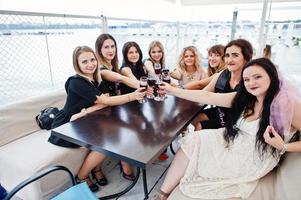 Group of 8 girls wear on black and 2 brides at hen party sitting at table and drinking rose champagne from glasses. photo