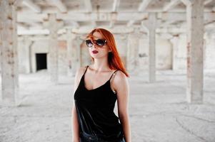 chica elegante de pelo rojo con gafas de sol vestidas de negro, contra un lugar abandonado. foto