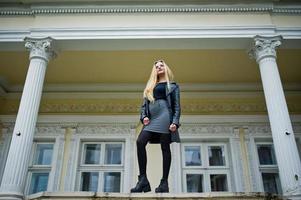 Blonde fashionable girl in long black leather coat posed against vintage house. photo