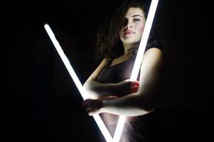 Curly brunette plus size model with two long led lamp at black room. photo