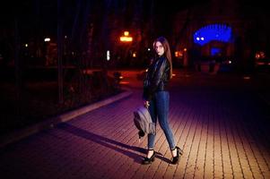 Night portrait of girl model wear on glasses, jeans and leather jacket, with backpack in hands, against blue lights garland of city street. photo