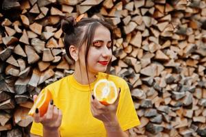 jovencita divertida con maquillaje brillante, vestida con camisa amarilla, sostenga una pieza de naranja sobre fondo de madera. foto