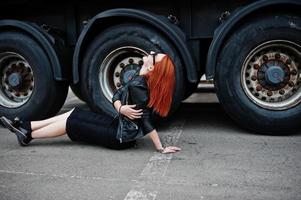 Red haired stylish girl wear in black, sitting against large truck wheels. photo