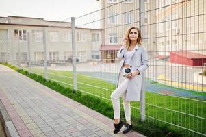 Stylish curly blonde model girl wear on white posing against fence. photo