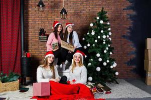 Four cute friends girls wear on warm sweaters, black pants and santa hats against new year tree with christmas decoration at studio. photo