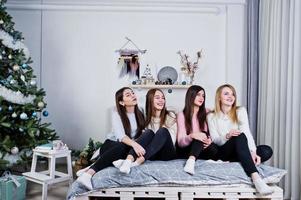 Four cute friends girls wear on warm sweaters and black pants on bed at new year decorated room on studio. photo