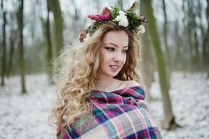 Curly cute blonde girl with wreath in checkered plaid at snowy forest in winter day. photo