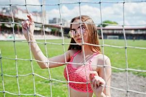 Fitness sporty girl in sportswear and sunglasses posed at stadium football gates outdoor sports. Happy sexy woman. photo