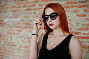 chica elegante de pelo rojo con gafas de sol vestidas de negro, contra un lugar abandonado con paredes de ladrillo. foto