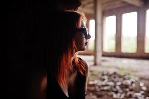 chica elegante de pelo rojo con gafas de sol vestidas de negro, contra un lugar abandonado con paredes de ladrillo. foto