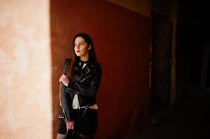 Stylish brunette girl wear on leather jacket and shorts with backpack against orange wall at shadows. photo