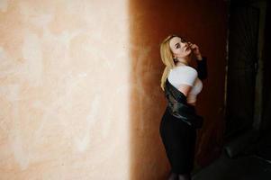 Elegant blonde girl wear on black leather jacket posing at streets of town background old wall with shadows. photo