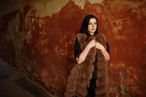 Fashion outdoor photo of gorgeous sensual woman with dark hair in elegant clothes and luxurious fur coat at old street with grunge walls.